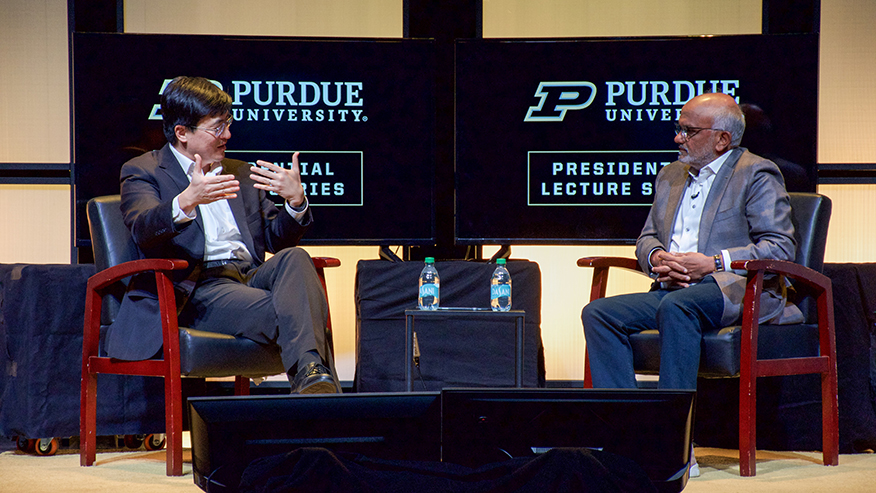 Two men sitting and talking on stage with tv screens behind them