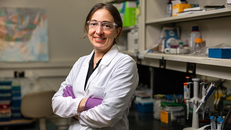 A smiling woman with brown hair and wearing a white lab coat, safety glasses and purple latex gloves stands with arms crossed in a laboratory.