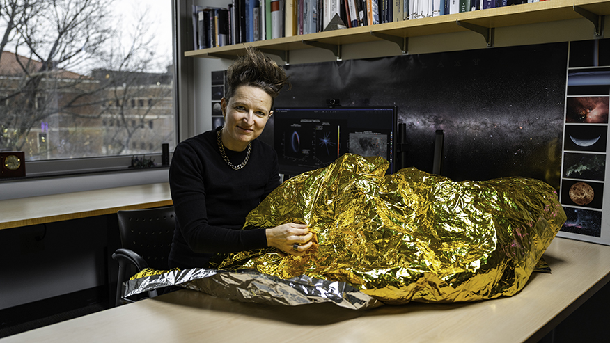 Purdue engineer Carolin Frueh holds a large sheet of metallic, golden material while sitting at a desk in her office