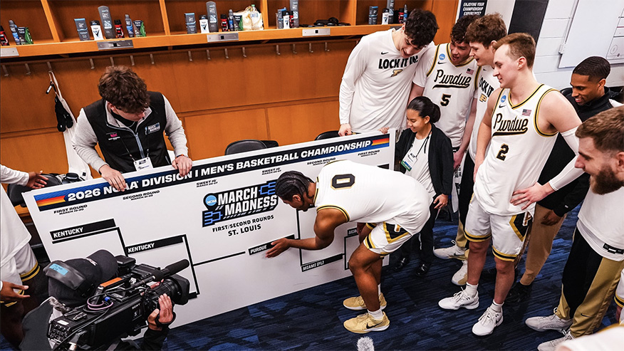 Purdue basketball players gather around an NCAA Men’s Basketball Championship bracket in a locker room to advance their name on bracket