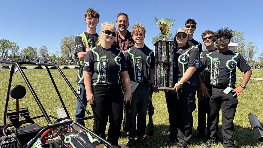 A group of people stand together by a kart with one person holding a large trophy