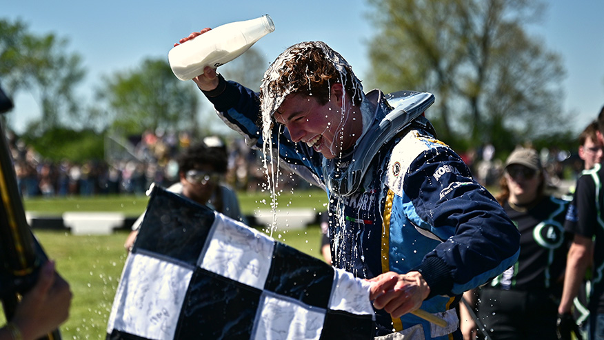 A man dressed in racing gear pours milk over his head and holds a checkered flag