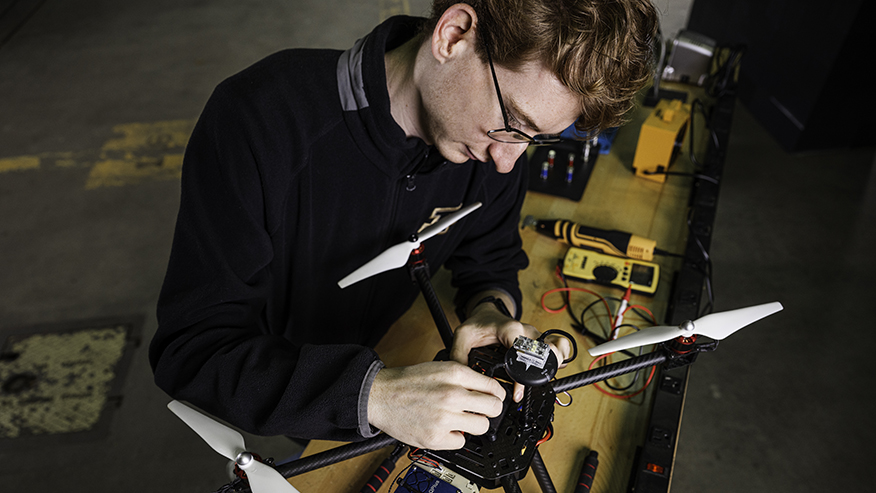 A Purdue student wearing glasses stands at a workstation, using both hands to adjust electronics on a quadcopter drone.