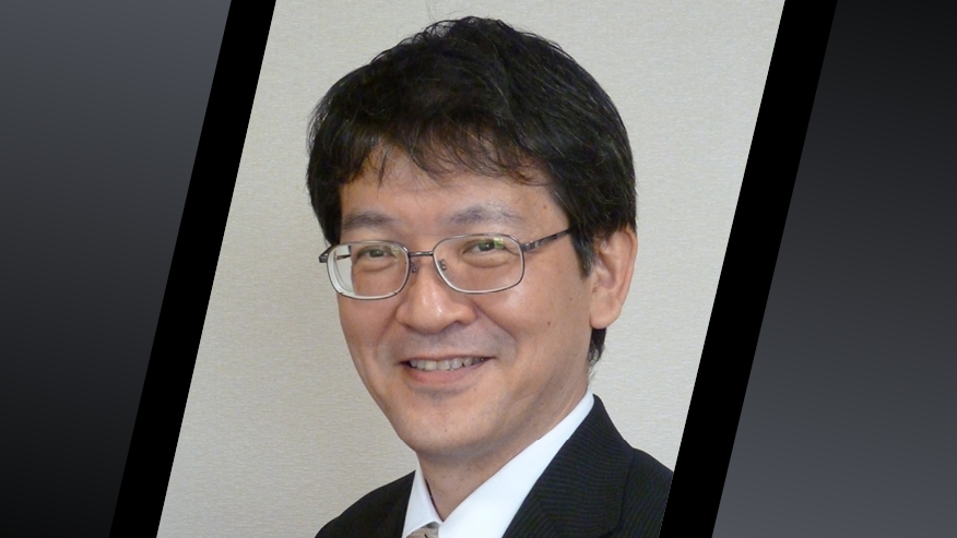 Portrait of a person wearing glasses and a dark suit, photographed against a light-colored background.