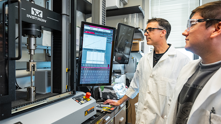 Two men in safety goggles look at a computer screen in a lab