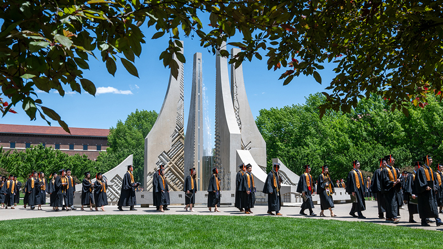 Students in graduation regalia walk in front of a scenic fountain, framed by vibrant trees in the foreground.