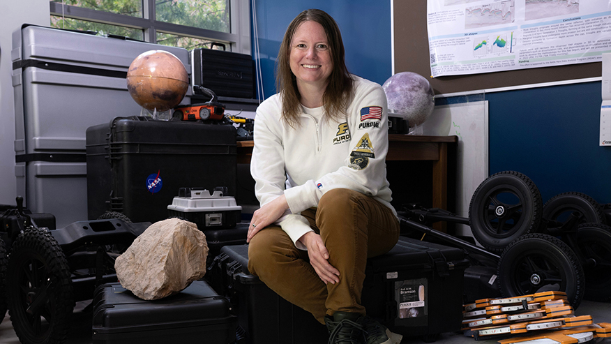 Ali Bramson seated among planetary models, a rock sample, rover wheels and equipment cases in a planetary science lab.
