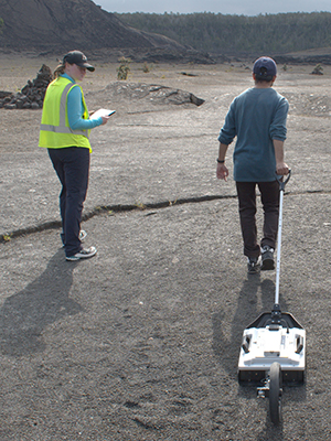 A researcher pulls ground-scanning equipment in a volcanic plain while Ali Bramson records notes.