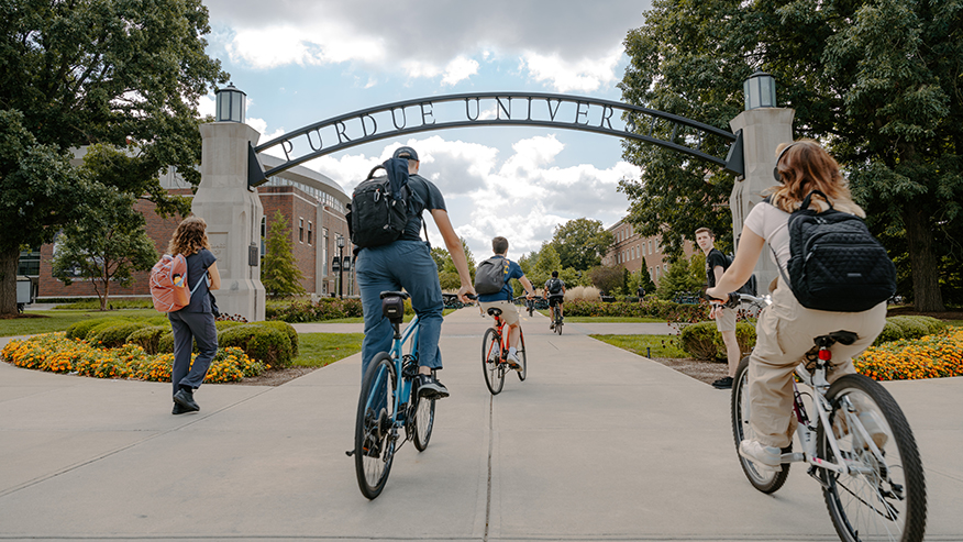 Students ride bicycles and walk beneath the Gateway to the Future arch at Purdue University.