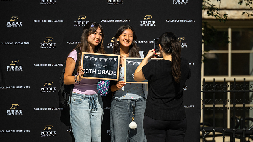Two young individuals pose with chalkboard signs marking their first day of 13th grade in front of a Purdue University backdrop, while another person captures the moment with a smartphone.