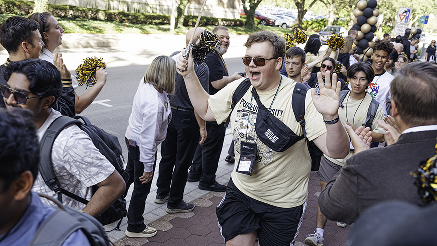 A smiling student in Purdue gear waves while walking through a cheering crowd at Boiler Gold Rush.