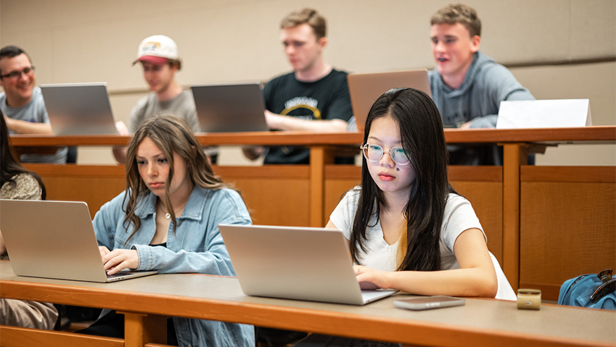 Students use laptops in a tiered classroom or lecture hall.