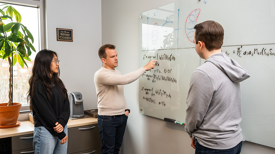Keith LeGrand talks with students and points to equations on a dry-erase board.