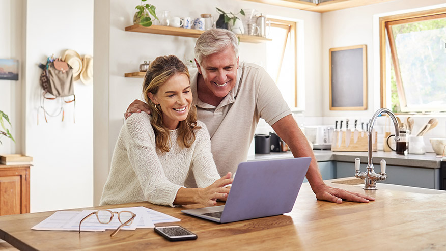 Two adults using a laptop together at a kitchen table, with papers, eyeglasses and a smartphone nearby.