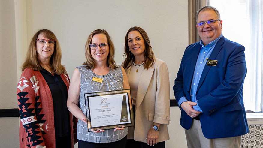 Four people pose indoors during an award presentation, with one person holding a framed certificate.