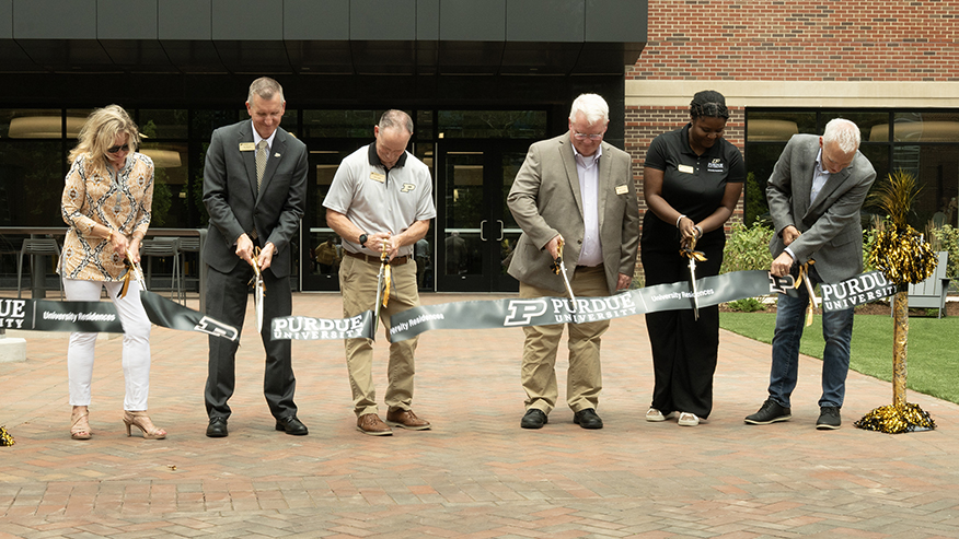 Six people holding large ceremonial scissors while cutting a ribbon outdoors in front of Purdue University’s South Hall.