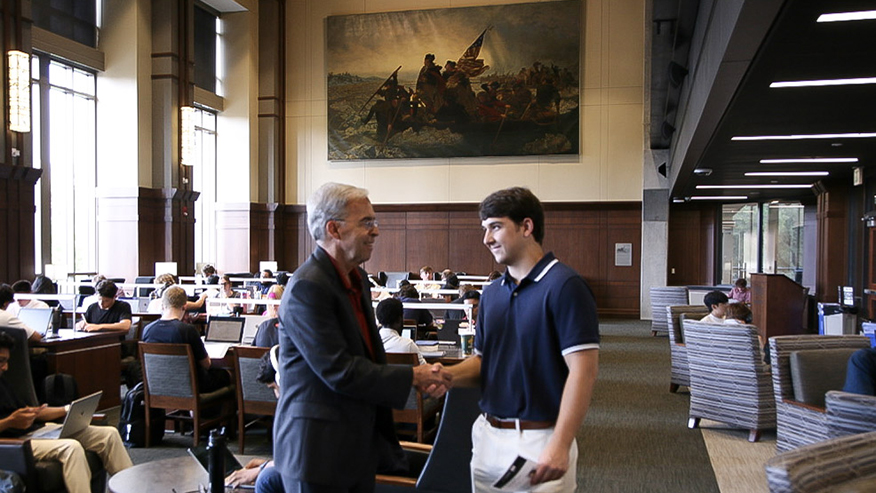 Two people shake hands in a large study and reading space, with a large historical painting hanging on the wall behind them. Students seated at tables work on laptops throughout the room.