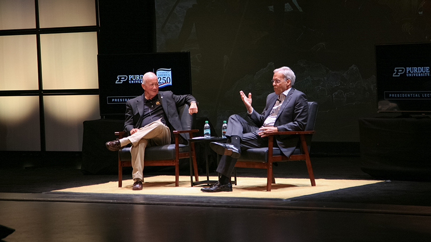 Two men sit on a stage and speak during a public discussion.
