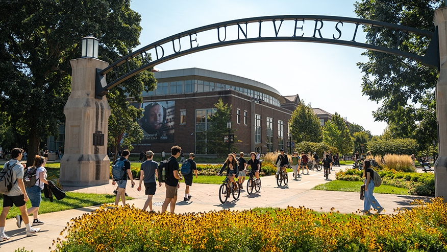 Students walk and cycle under a “Purdue University” archway on a sunny day, with a modern building and vibrant yellow flowers in the foreground.