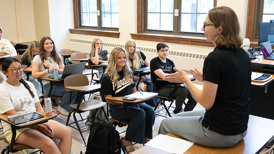 An instructor in a classroom engages with students seated at desks, some using laptops and tablets, while others listen attentively.