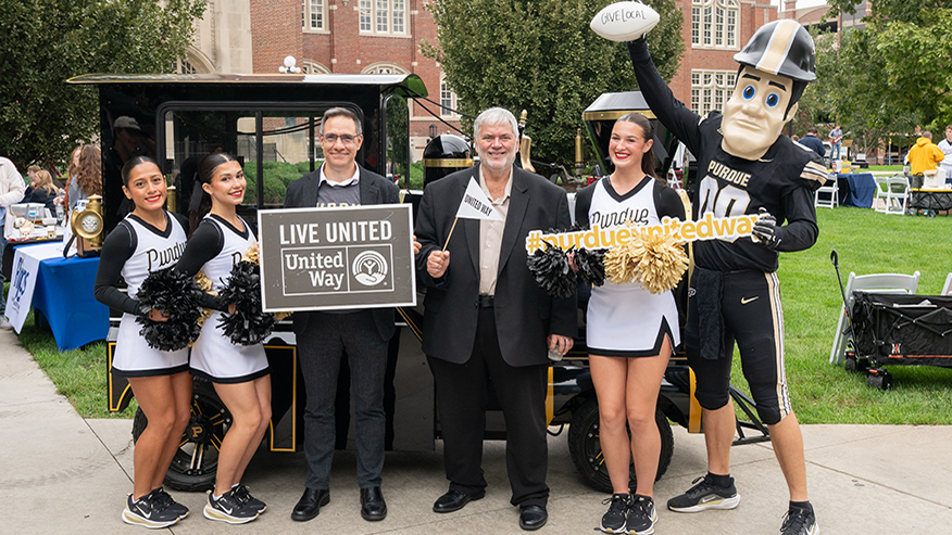Two people stand with cheerleaders and Purdue Pete near the Boilermaker Xtra Special.