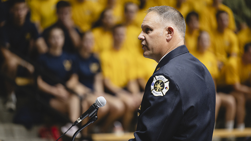 Man in fire uniform speaks behind microphone with blurred group in background