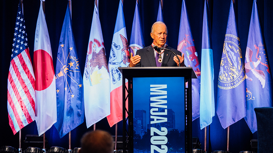 Purdue President Mitch Daniels speaks at a podium during the Midwest U.S.-Japan Association Conference, with U.S., Japanese, Indiana and other flags displayed behind him.