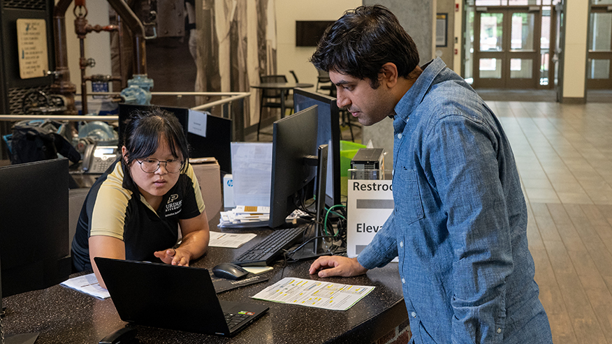 Person at a reception desk looks at a laptop with a college student.