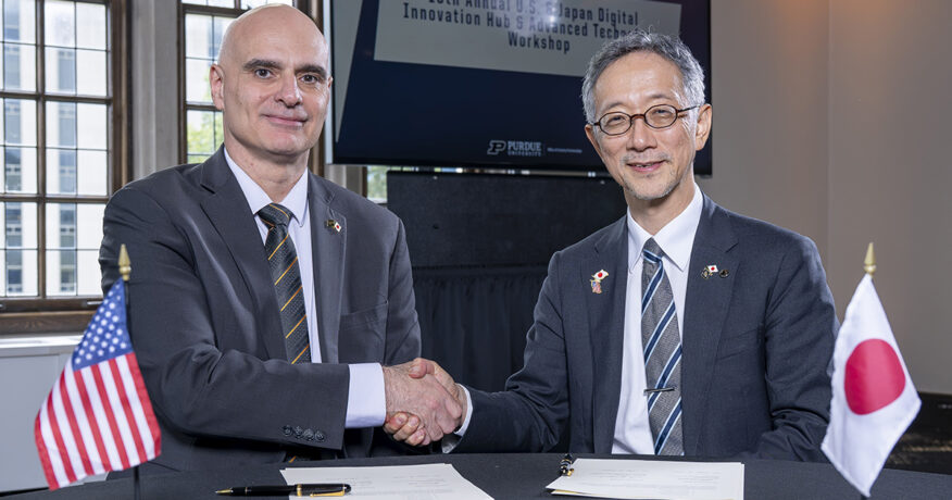 Two people — Dimitrios Peroulis of Purdue and Masahiro Yamaguchi of Tohoku University — shake hands after signing a letter of intent, flanked by the American and Japanese flags.