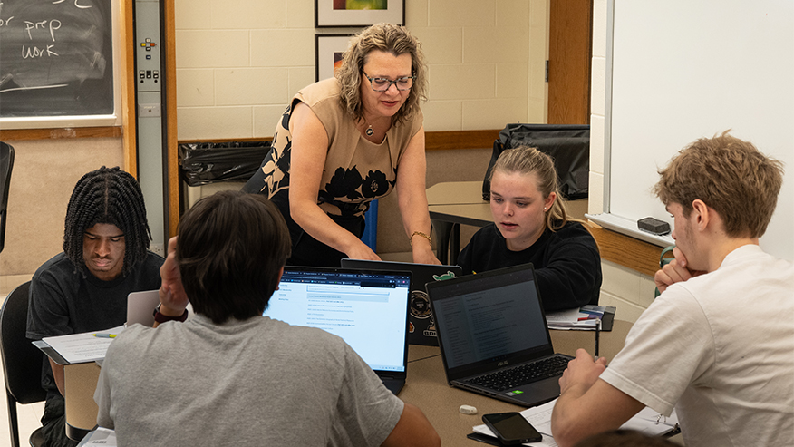 A faculty member works with students in a classroom discussion using laptops and course materials.
