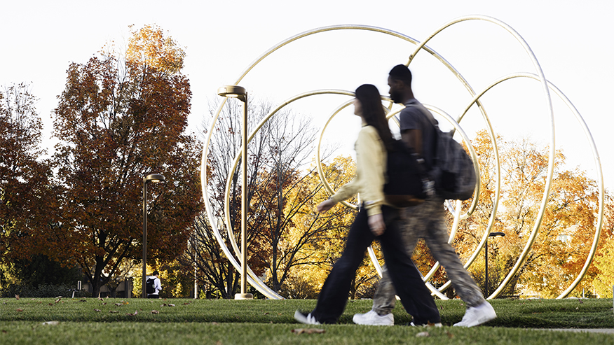Students walk past a circular art piece with fall trees in the background.