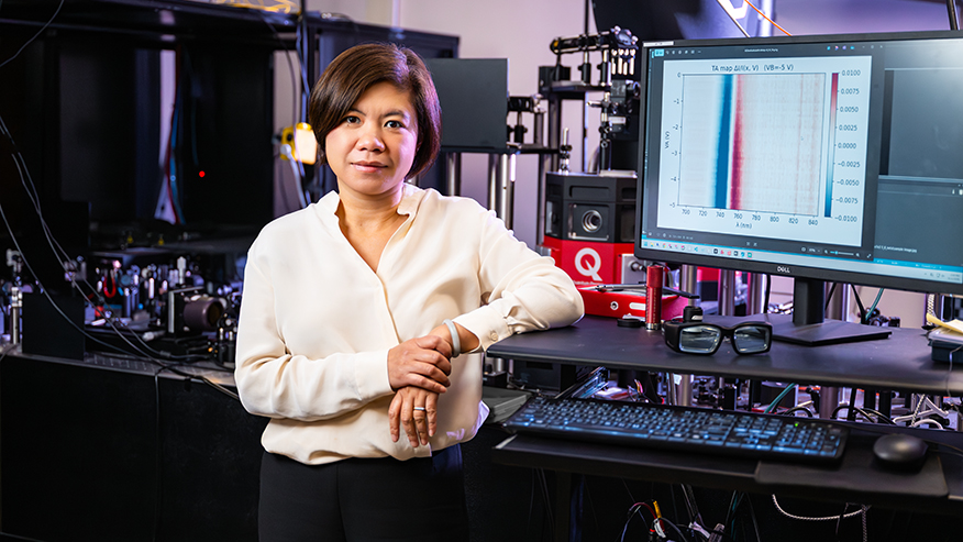 A woman in a white blouse and black pants stands next to a computer monitor in a lab equipped with optical and laser equipment.