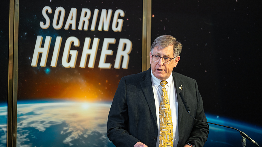 A person in a suit speaks at podium before a space-themed backdrop showing Earth and the words “Soaring higher.”