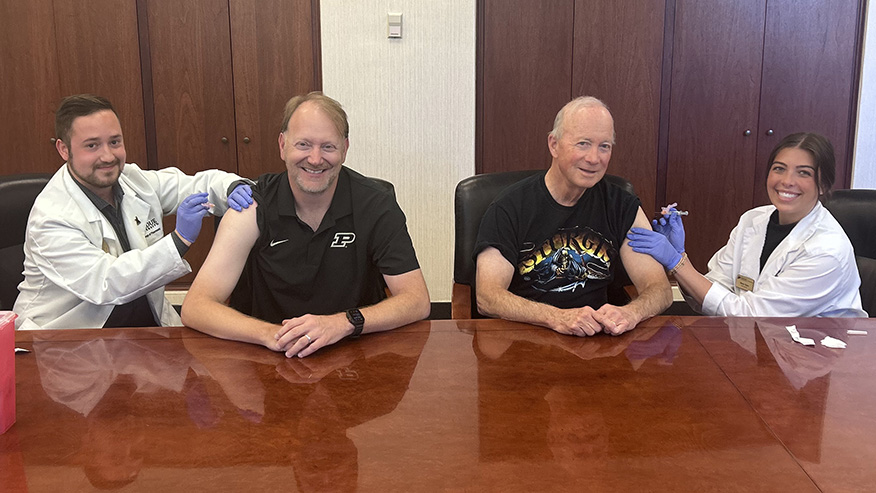 Purdue’s provost and president receive vaccinations at a conference table.