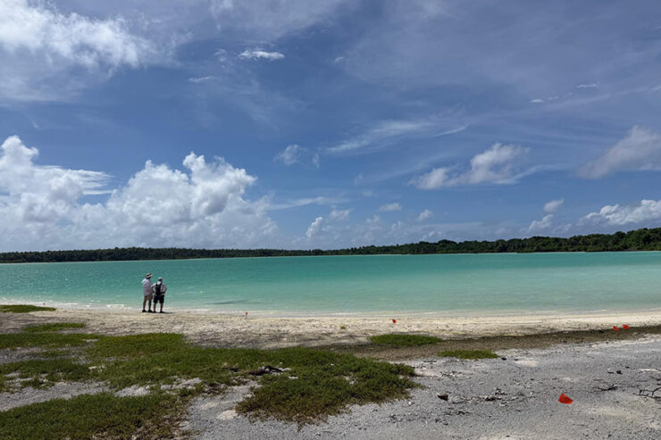 Two expedition members stand along the shore of Nikumaroro’s lagoon overlooking turquoise water.