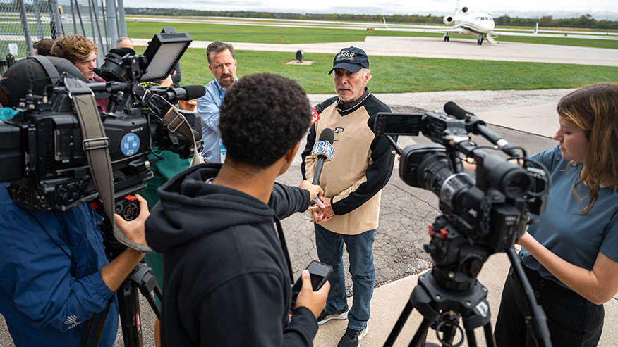 A person in a cap and vest is being interviewed by reporters with cameras and microphones on an airfield, with a private jet visible in the background.