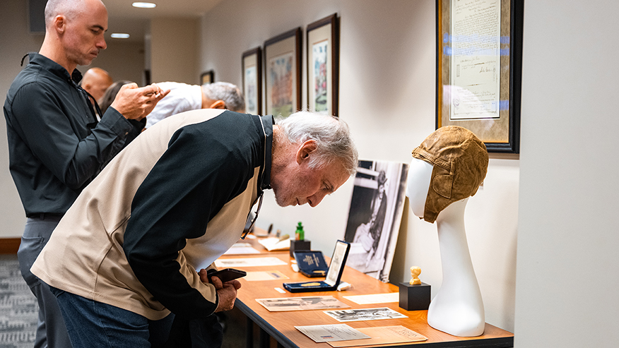 A person leans closely over a display table examining historical artifacts, including a hat on a mannequin and framed documents, while others observe nearby.