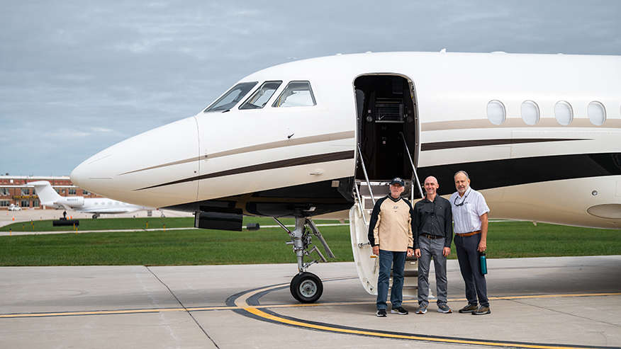 Three individuals stand on the tarmac beside a private jet with its door open, under an overcast sky.