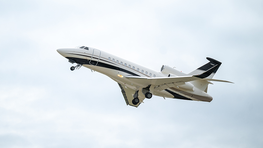 A white private jet with black accents is airborne, its landing gear extended against a cloudy sky.