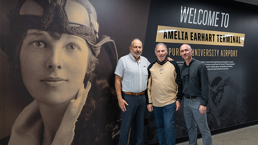 Three individuals stand in front of a large mural honoring Amelia Earhart at the Amelia Earhart Terminal, with signage indicating the location is Purdue University Airport.