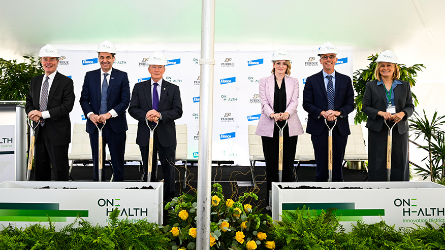 Six individuals in hard hats stand at a groundbreaking ceremony for the One Health Innovation District, each holding a shovel poised to break ground.