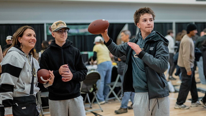 Three individuals stand in a gymnasium, with one person holding a football aloft while another holds a football in their hands.
