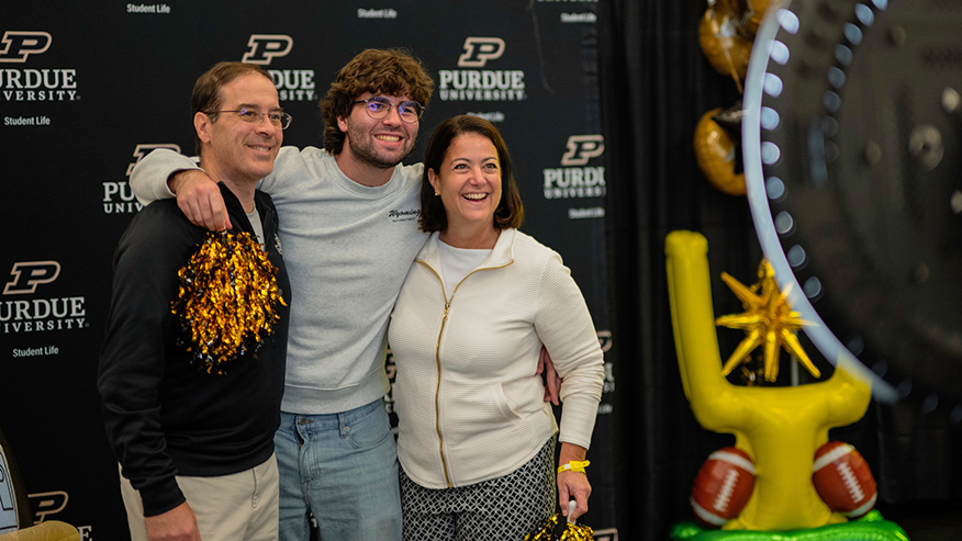 Three people pose together in front of a Purdue University backdrop, with one holding gold pom-poms and another standing beside a large yellow football goalpost decoration.