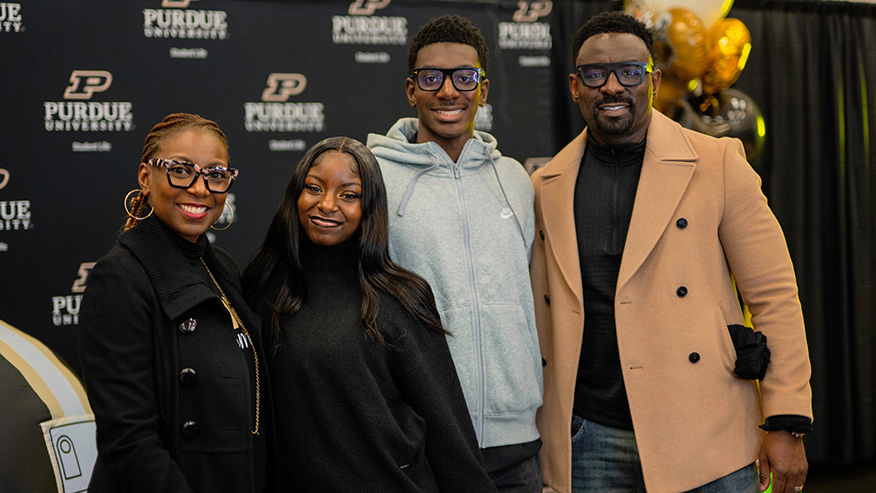 Four individuals pose together in front of a Purdue University backdrop, smiling for the camera with festive balloons visible in the background.