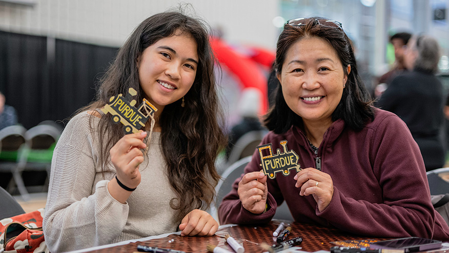 Two individuals smile while holding handmade Purdue-themed train-shaped crafts at a table with crafting supplies.