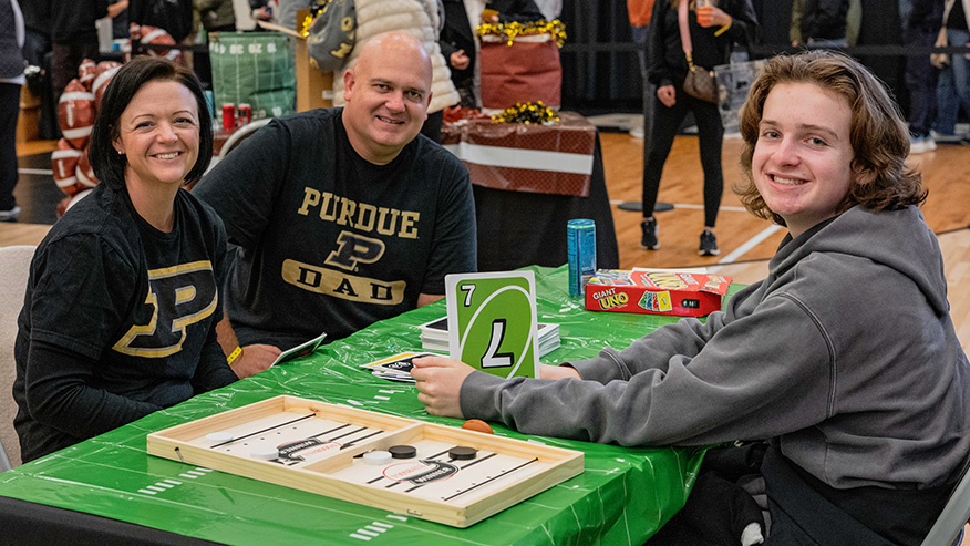 Three people wearing Purdue apparel smile while playing games at a table covered with a green tablecloth.