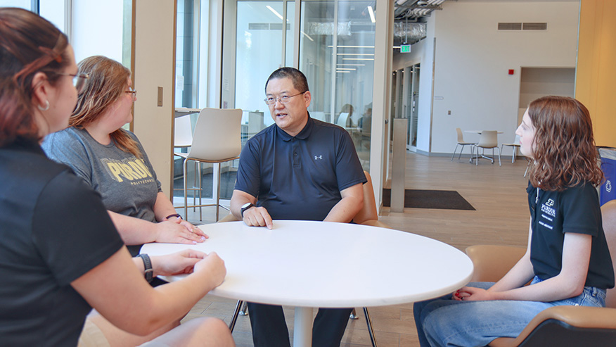 A man talks with three students around a table in the lobby of an academic building.