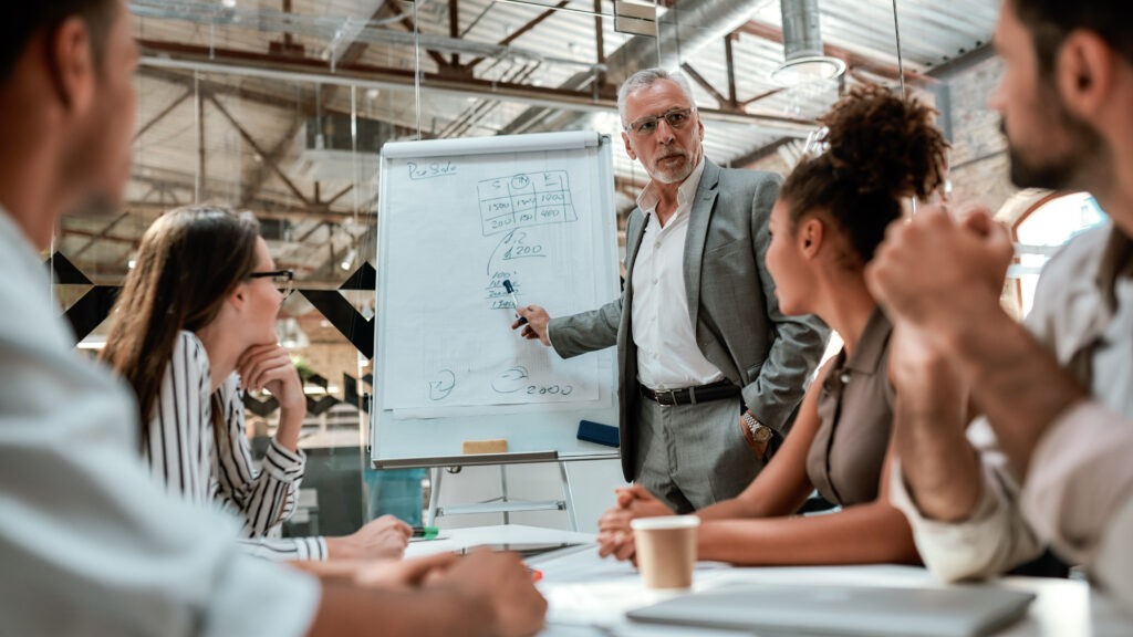 Business team. Confident mature businessman standing near whiteboard and explaining something to his young colleagues during meeting in the office. Teamwork. Presentation