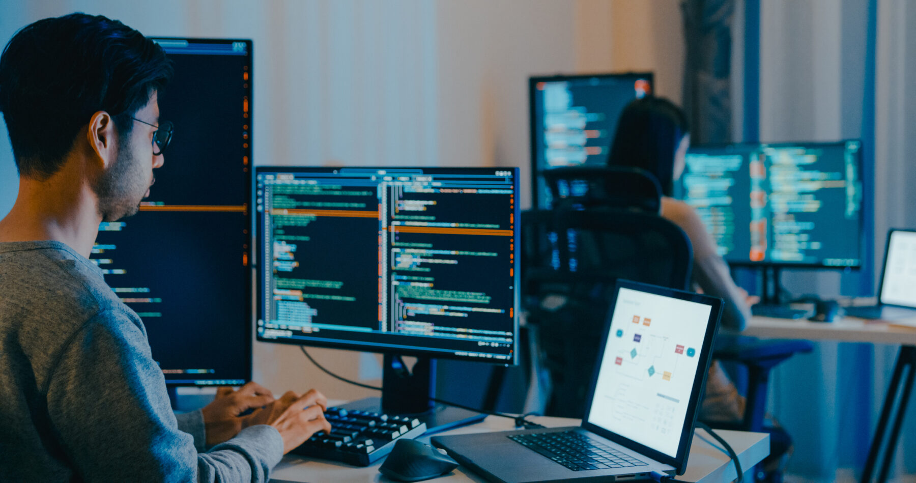 A man sits at a desk typing on a laptop. He is surrounded by monitors that show data analyses.