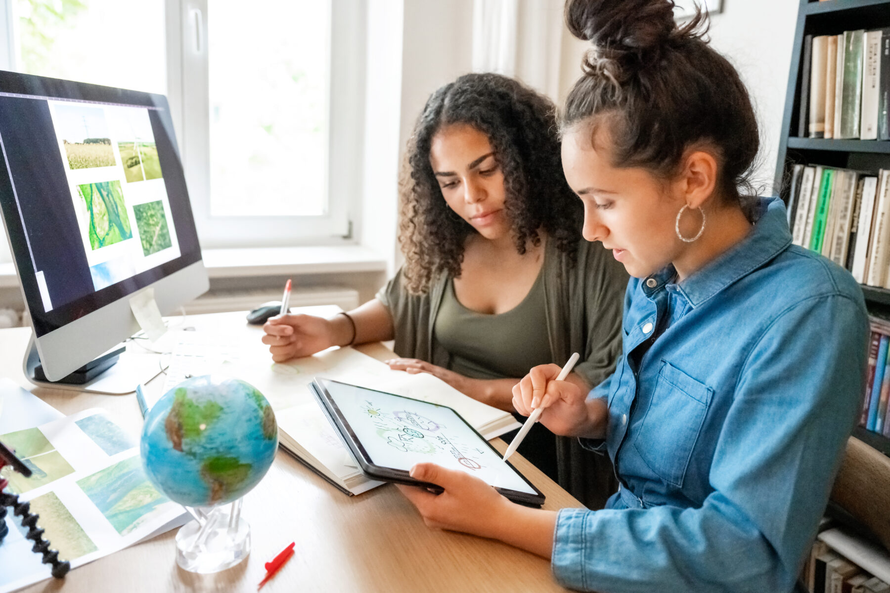 Two young women sit at a desk and map out a lesson plan using a notebook and computer.